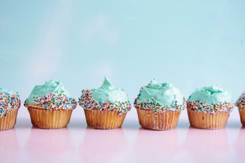 Six vanilla cupcakes with light blue frosting and sprinkles lined up against a blue background.